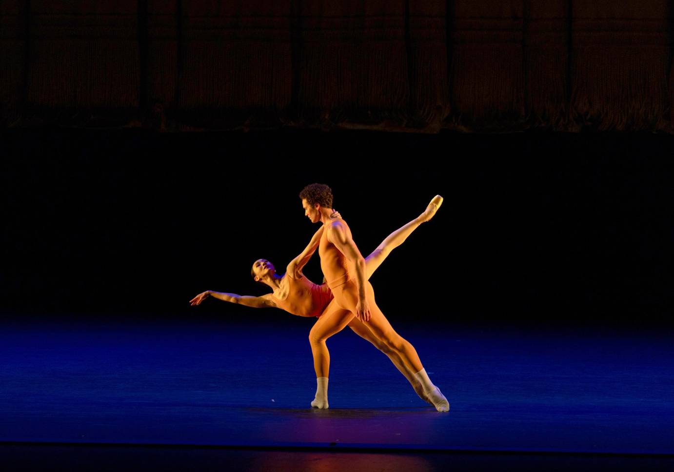 a duet between a male and female partner..both wear bright orange unitards, and the stage floor is colored a striking complementary blue. The man , in a forward lunge, stands above the woman, as she tilts forward on pointe, in an unusual balance, with her grabbing his neck as her body is pitched almost parallel to the floor 