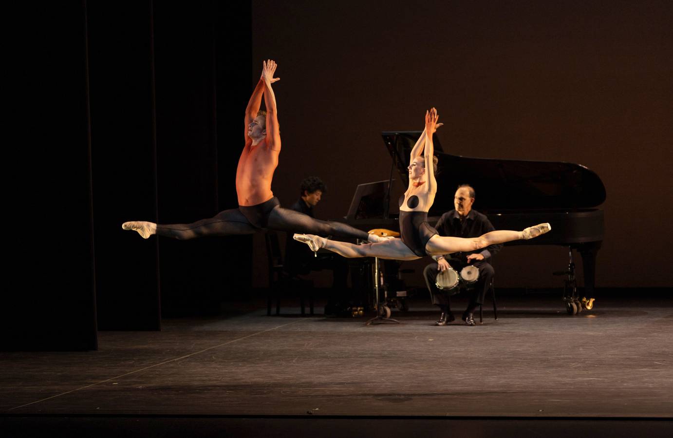 a pianist and drummer are on stage playing away as a male and female dancer hover high in the air , both in identical split leaps.