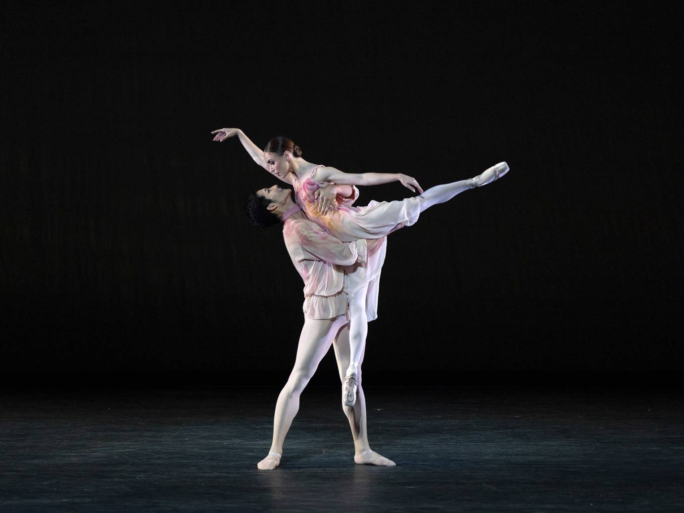 2 dancers in pale costumes lightly colored with pinks reds and orange watercolor designs  that flow into eachother. Similarly the woman flows into her male partner as he holds her in front of him while she soars in a high arabesque. 