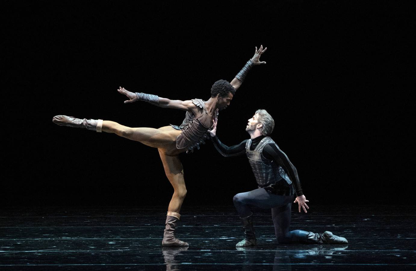 A man in arabesque arms stretched wide looms over another man kneeling at his feet. The kneeling man's hand presses on the other man's chest.