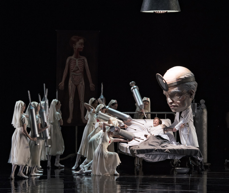 A group of women in white nurse costumes hold oversized prop syringes. Daniel Simkin sits in a hospital bed awaiting a shot. Another dancers wheres an oversized doctor's mask and gets ready to administer the shot.