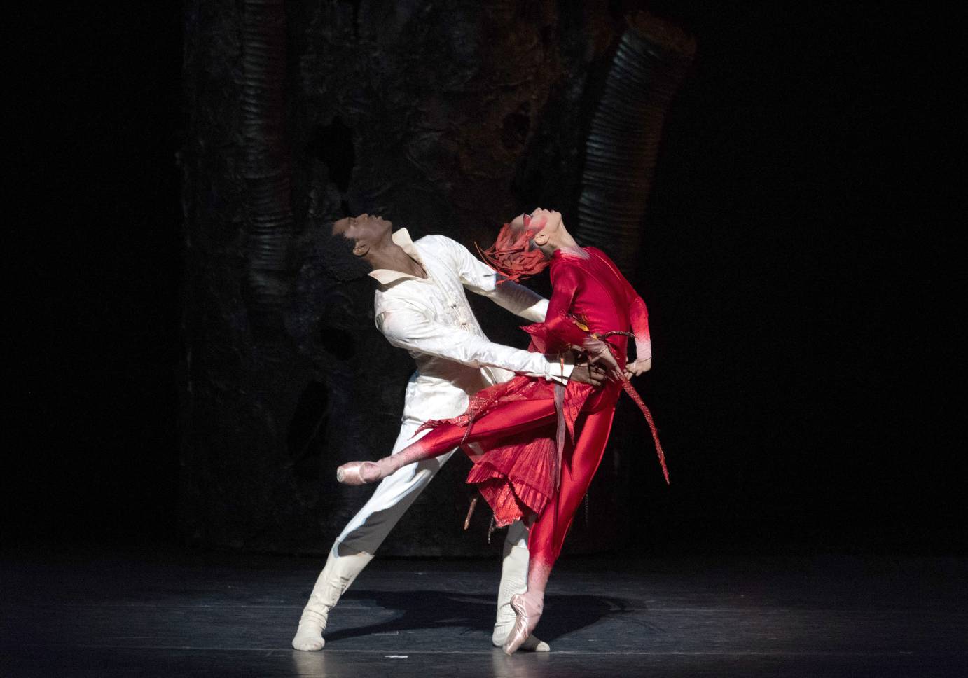 A ballerina dressed in red and her partner costumed in white both look upward as he holds her hips.