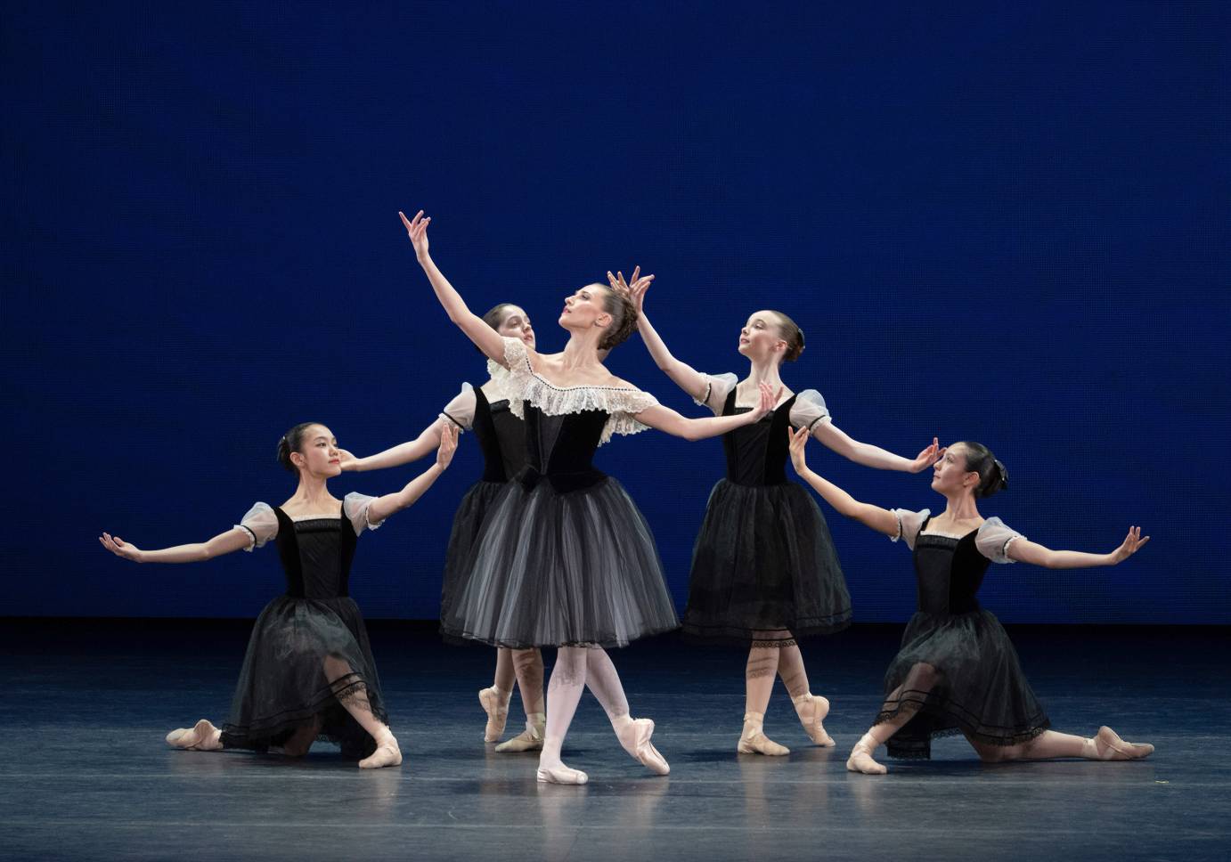 A ballerina in a black flowing dress stands with her right arm and gaze upward. Four ballerinas kneel and stand behind her, their gazes and arms following hers.