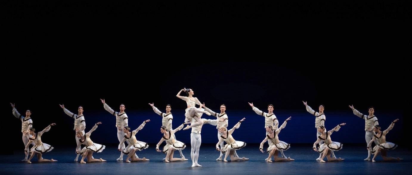 A male ballet dancer lifts a ballerina onto his shoulder, both looking stage left. Eight couples are behind them; the ballerinas kneeling and looking diagonally down while their partners are looking outward with right arm lifted on the diagonal.