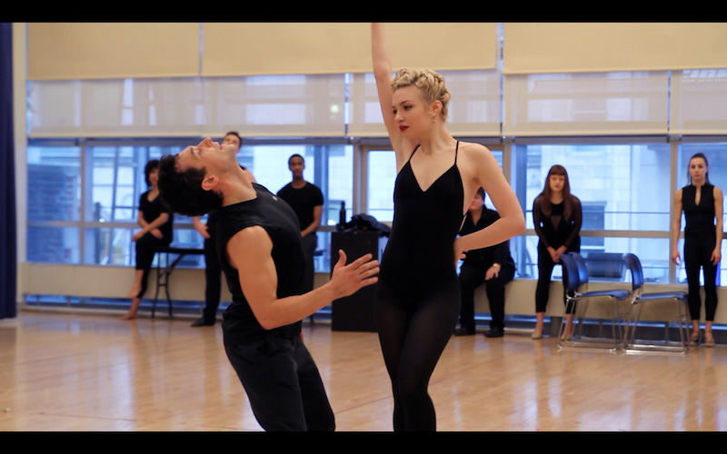 A man and woman dance together. She, with her hand on her hip, while he leans back with his head facing the ceiling. Dancers watch near the windows