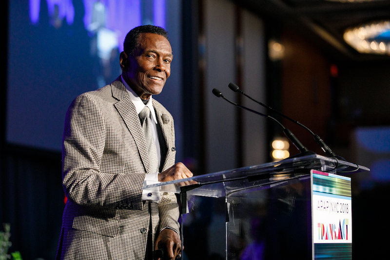 Arthur Mitchell, looking dapper in a plaid suit smiles from the camera as he stands at a podium.