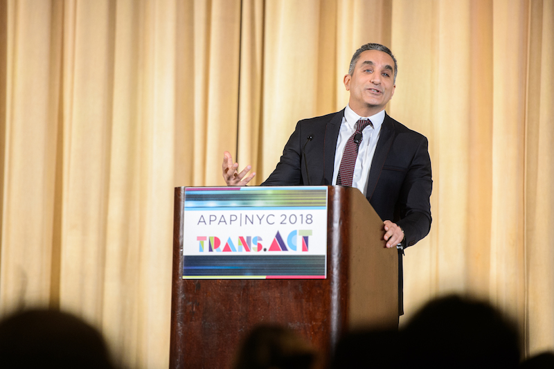 Bassem Youssef in a suit and tie stands at a podium in front of a gold curtain to address an audience. 