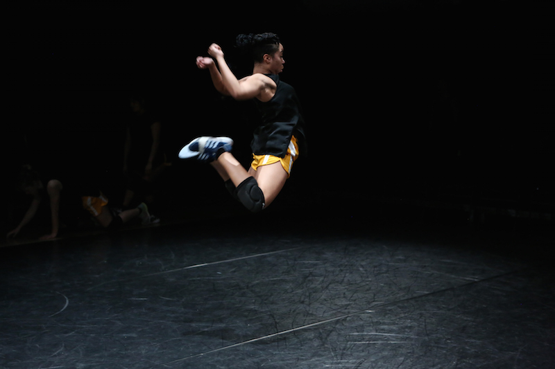 A woman jumps into the air kicking her legs behind her seat and arms swing to her sides. The view is in profile. She wears a black tank top, orange athletic shorts, knee pads and sneakers.