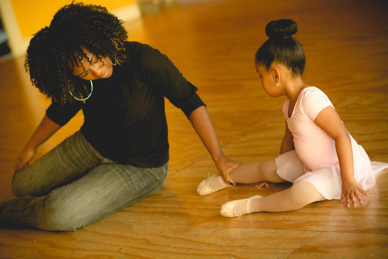 Karisma sits on the floor next to one of her young students. She touches her ankle.