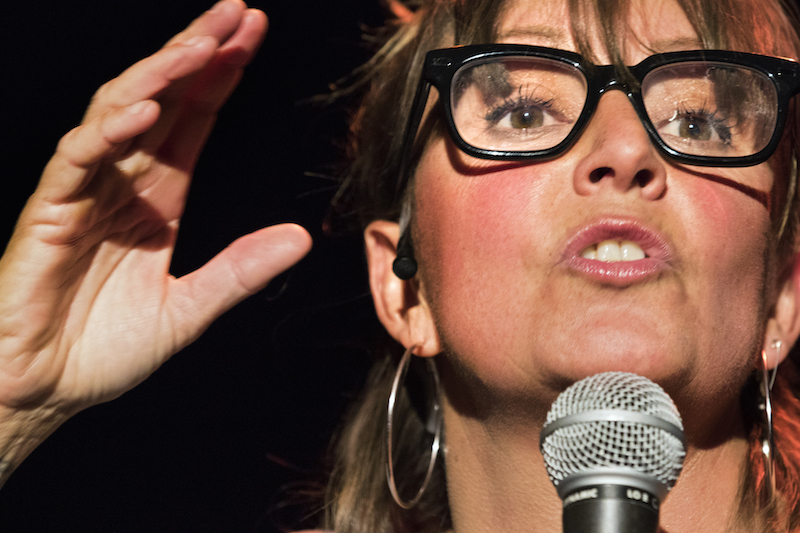 Wearing nerd glases, Adrienne Truscott speaks into a microphone.