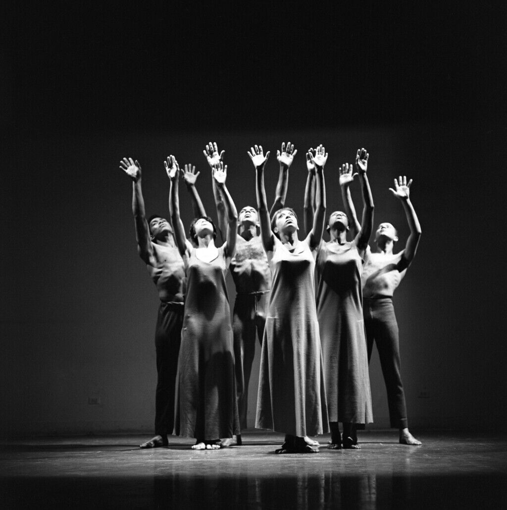 Six lithe women and men dancers facing the audience and dressed in simple ankle-length dresses reach reach their arms to the sky with palms outward.