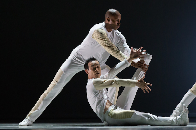 Two dancers in white. One sits on the floor with his legs straight and scissor-like. The other is in a wide stand behind him. Leaning towards his right bent leg.