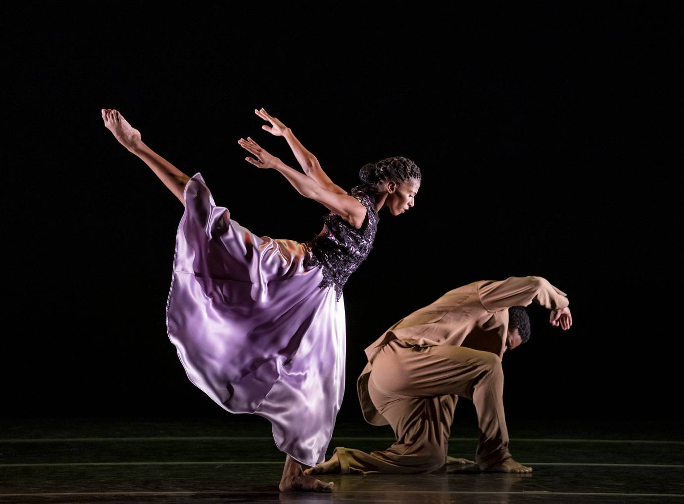 One man kneels while a woman in a purple skirt jumps with one leg high 