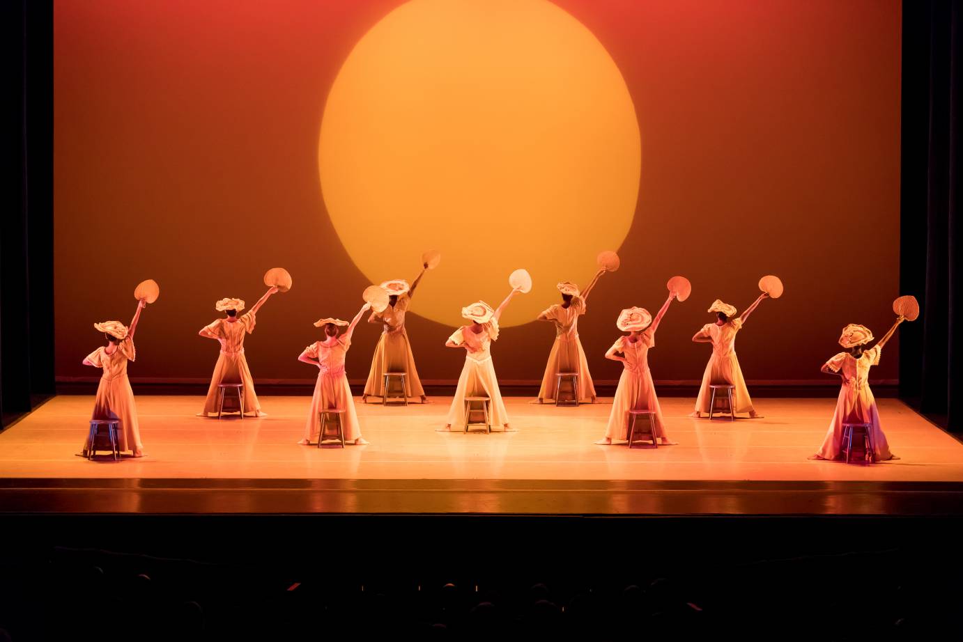 Against a backdrop of a sun, woman in hats hold up fans