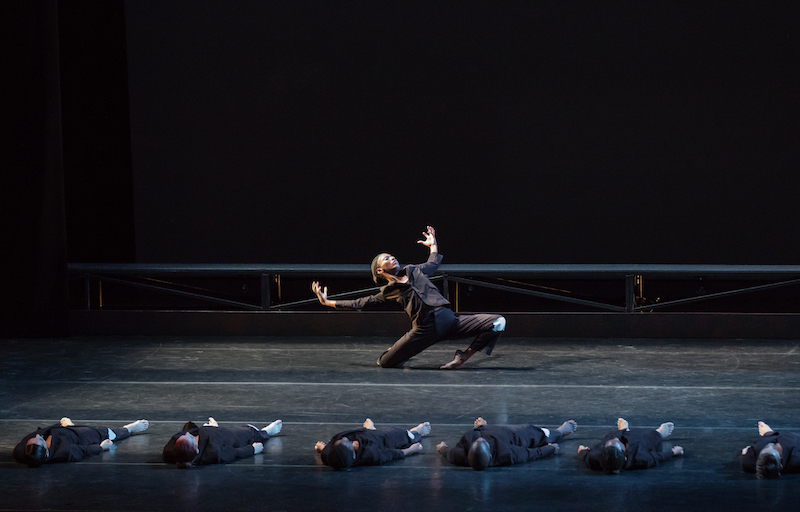 Dancers in black suit lay down in a line while a female dancer splay her arms and rolls over her foot as she descends to the ground.