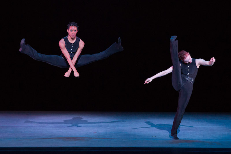 One woman jumps high into the air; her legs splay parallel to the ground. Another woman kicks her leg close to her head.