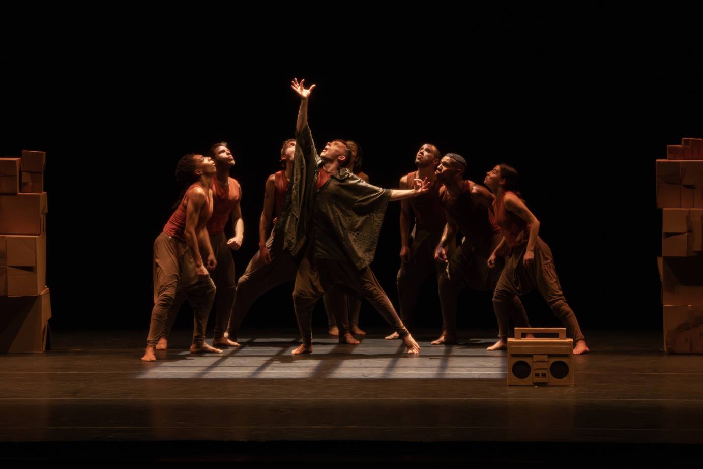 dancers, all wearing versions of orange tops and grey bottoms surround a central figure who is reaching up toward a light. The group is flanked by cardboard boxes. and in front of them is a boom box made of cardboard.