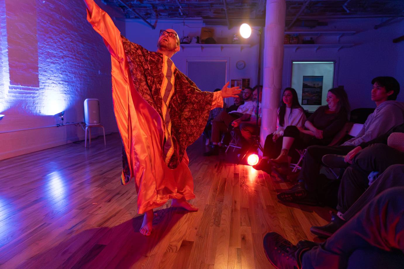 A bald headed bespeckled performer, dressed in a full length orange liturgical vestments, reaches upward on the diagonal