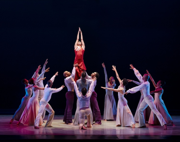 the company lifts a woman in red by the legs as her hands, clasped together, reach towards the sky. Those who aren't lifting her open their arms to her in respect and admiration 