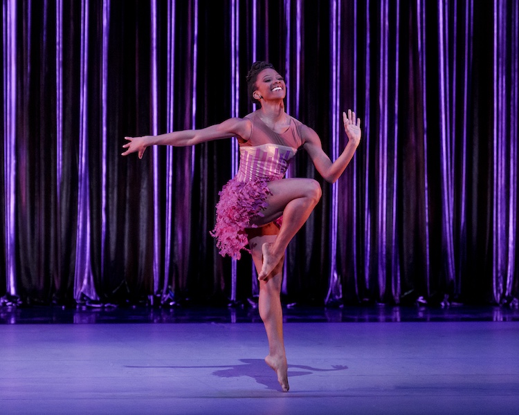 a bubbly smiling female dancer in a tight top and feathery light purple short skirt appears to be mid Charleston as she jigs and smiles before a shiny purple curtain