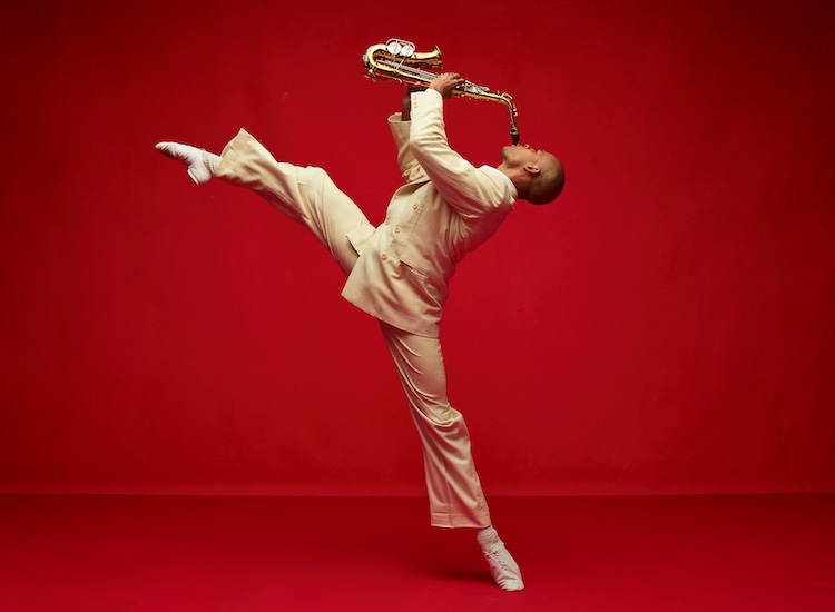 against a red wall and floor a man in a cream colored suit gloriously balances on one leg as he leans backward and mimes playing a saxophone. His other leg high and fully energized in the air.