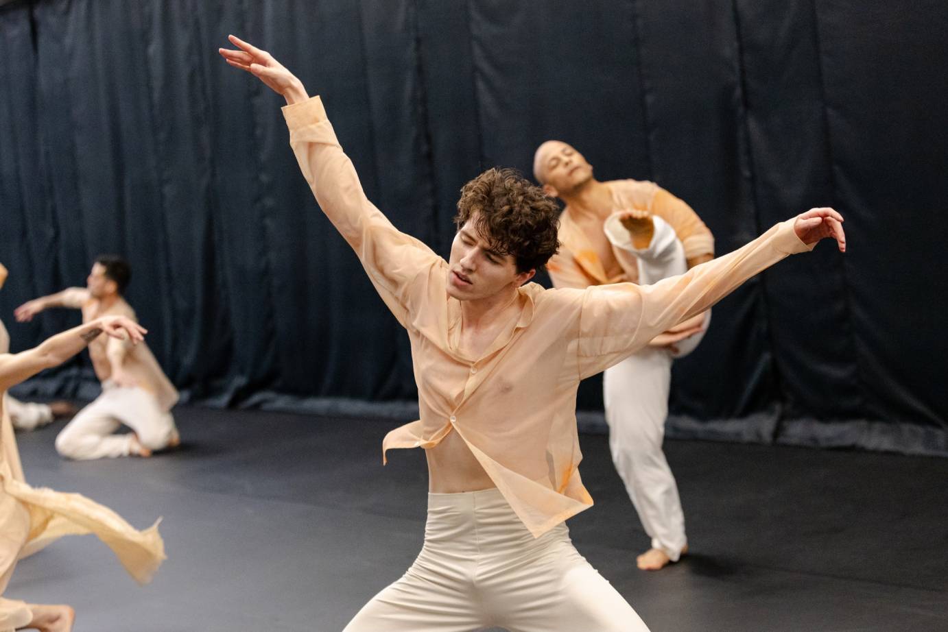 A man in a light orange shirt tilting to the right, his face soft, hair tosled, tosses both arms to the diagonal. A man behind tilts the other direction.