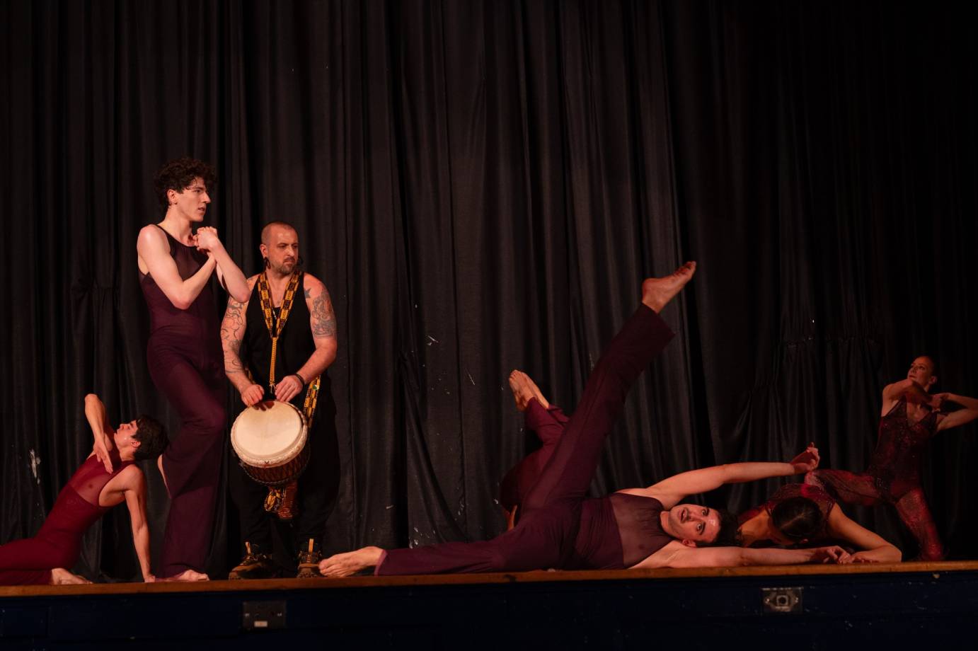 A tatooed drummer playing a small drum in front of his legs is upstage of six maroon clad dancers in various dance positions: standing with hands clasped to chest, kneeling with elbow high, on the side of the body with leg to ceiling and standing with both elbows jutting horizontally.