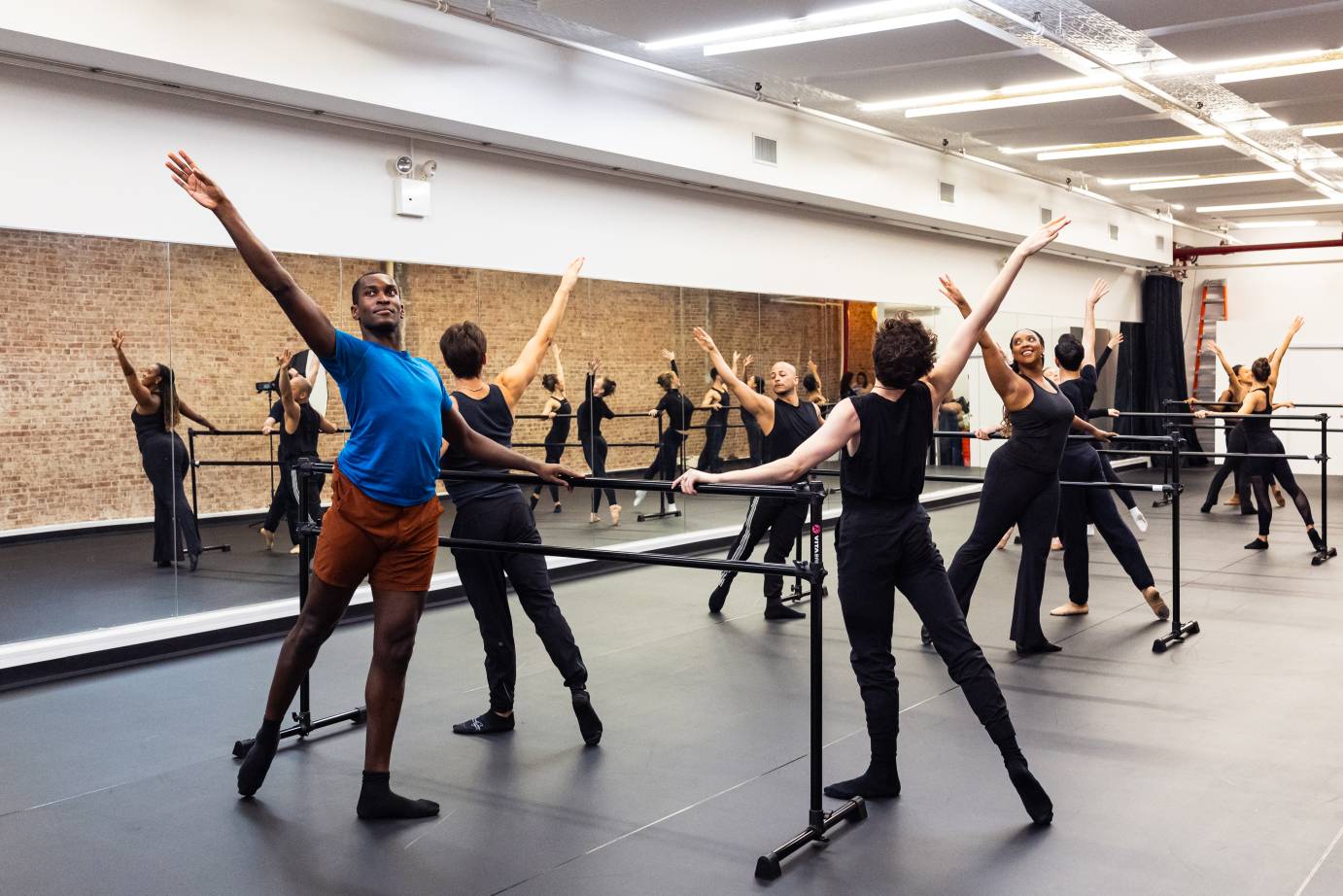 ASDC taking ballet class in the new studio: a mirrored wall reflects the other wall which is brick. Female and male company members are standing at black barres with the right leg tenduing back and the right arm extended on the diagonal.
