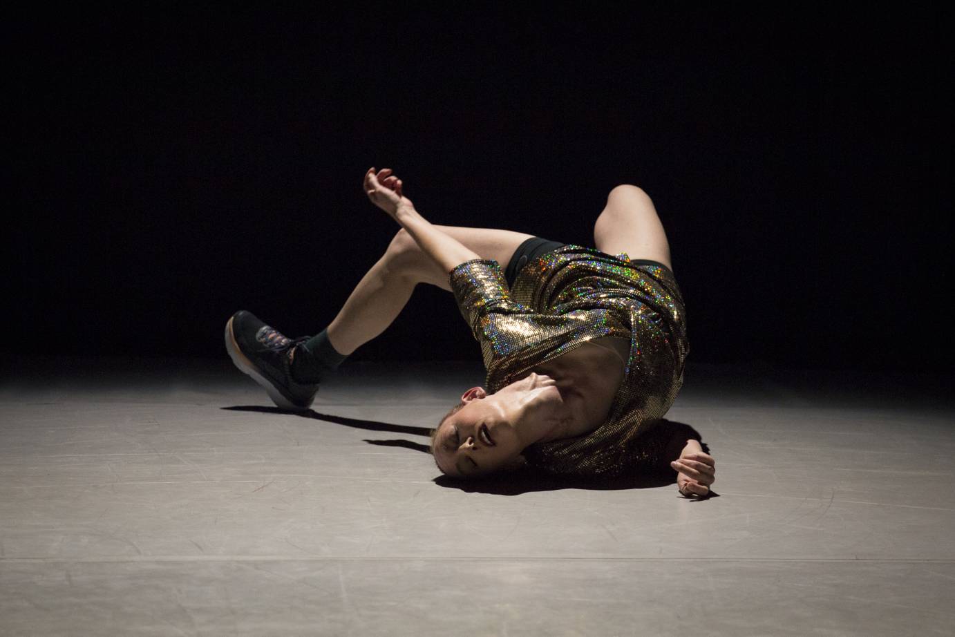 Woman curved and spiraled in an arch on the floor, she lies on her right shoulder with her face upside down. Her left black sneakerd foot is flexed. She wears a patterned, shiny top and short black shorts.
