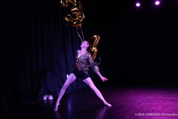 White woman with long legs and short black shorts, legs in a new lunge holds the ribbons to large gold helium balloons with her right hand behind her back. Her left arm hangs long parallel to her upright body. Whe wears a sheer black printed blouse