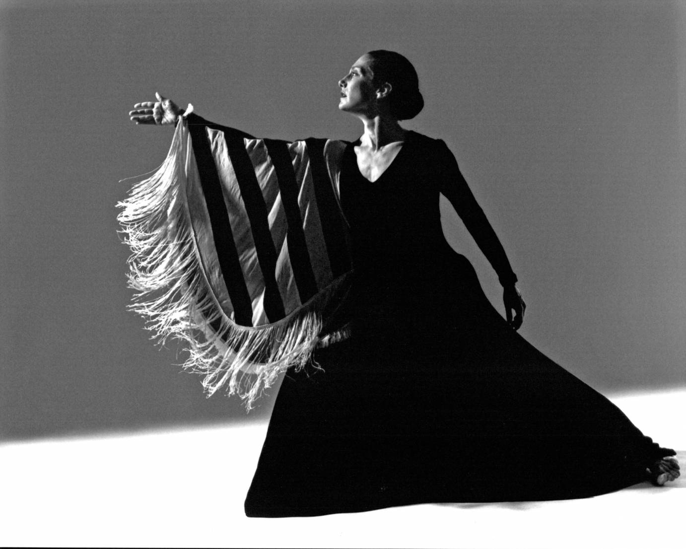 a black and white photograph of a woman in a dramatic lunging profile, her hair is in a tight bun, she wears a long black gown and a striped shawl hangs from her extended arm