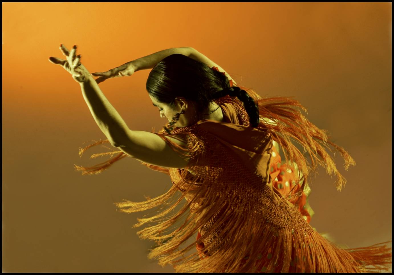 a very dynamically caught photo of a Spanish dancer from the back; she wears an orange fringed dress that swings in motion, and she is beautifully set against an orange background
