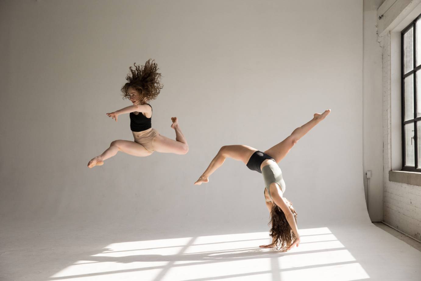 two women in midriff tops and beige briefs dance in a room against a white back drop... one is caught in mid-air her red curls flying above her, the other is in a handstand with her legs bent, you can feel their energy  