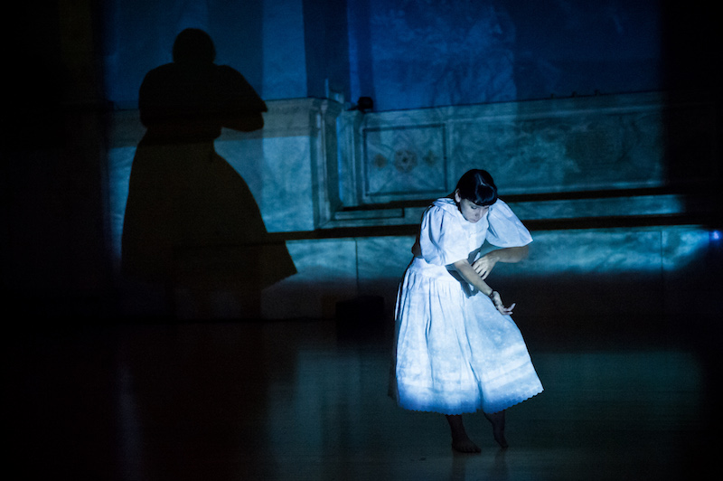 Rosy Simas in a white dress draws her fingers across her forearm in semi-darkness. Her shadow is on the back wall.