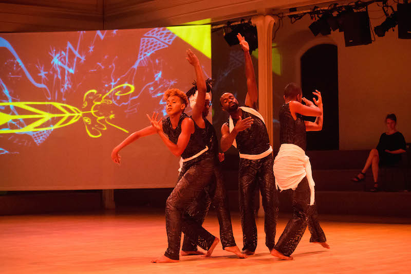 Four bodies dance in a clump. A projection of neon graphic is behind them.