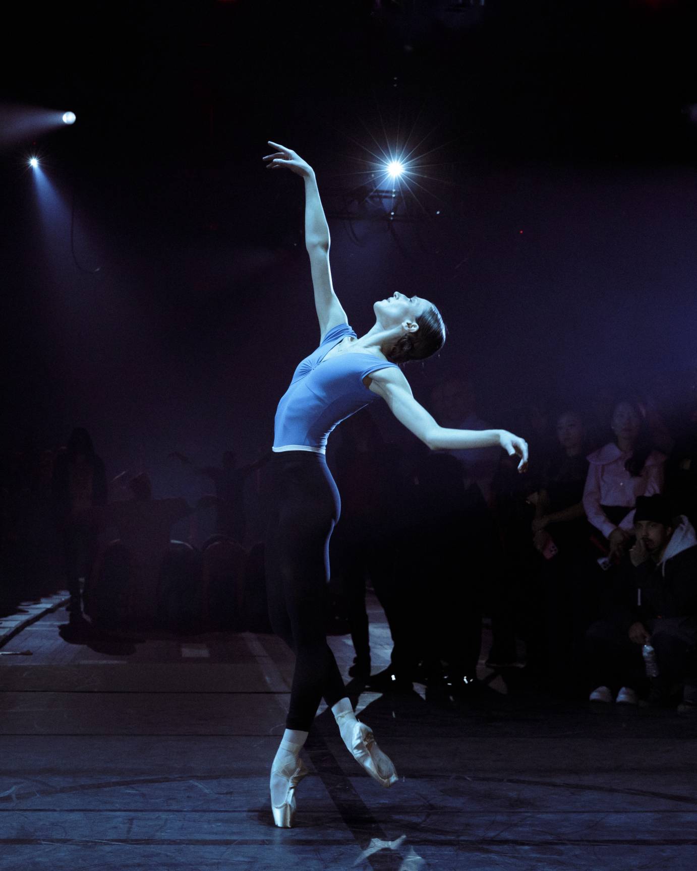 A ballerina on point, reaches one arm skyward as she tilts her head backward.
