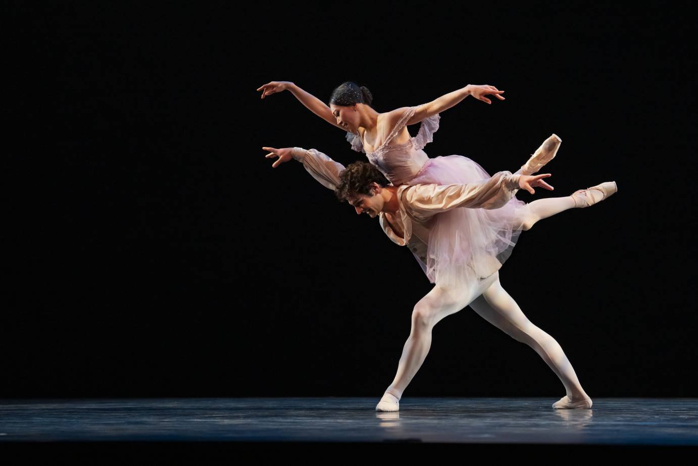 A ballerina dressed in a diaphanous pastel dress perches atop the upper back of a male ballet dancer in a tight lunge. Their faces look down as their arms wing to the sides pointer finger prominent