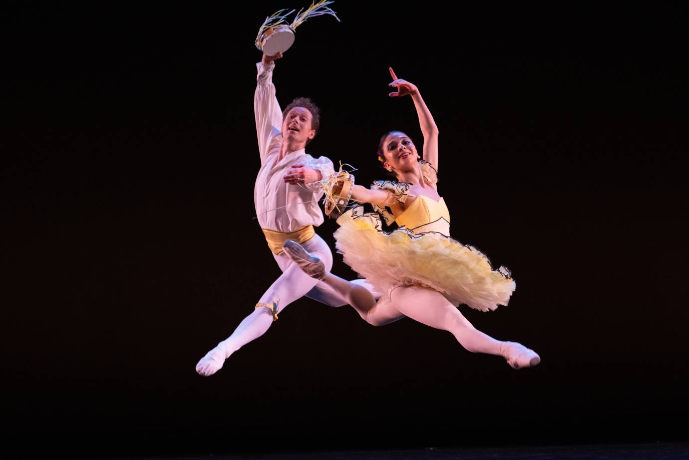 A male ballet dancer and a ballerina with backs to one another but front arms parallel to one another, leap toward downstage. He holds a tamborine aloft as she holds the tamborine extended toward downstage.