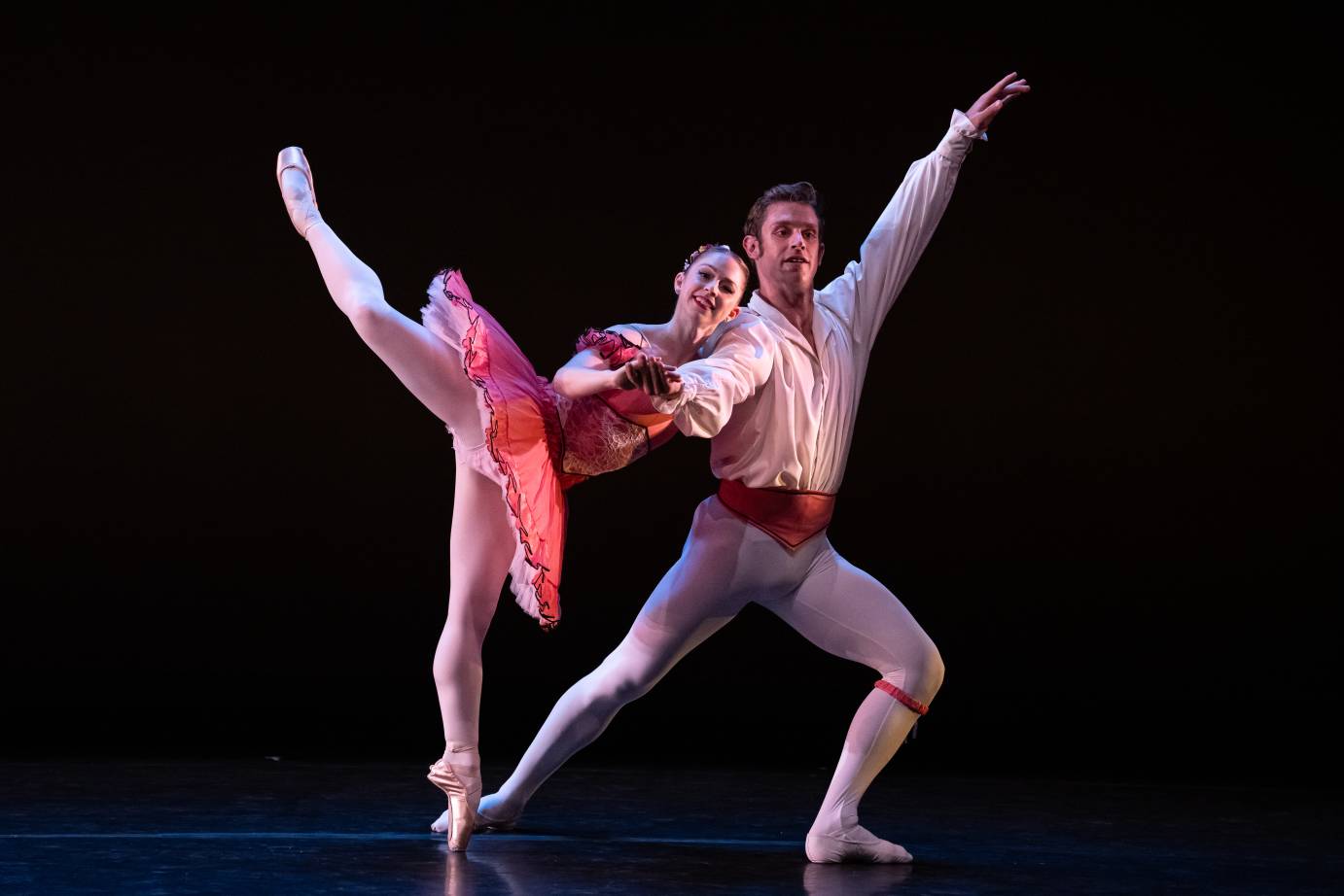 A smiling ballerina leans on the upper shoulder and arm of a male ballet dancer in a slight lunge. Both face forward while his free arm shoots to the ceiling equaled by her very high-reaching leg in attitude.