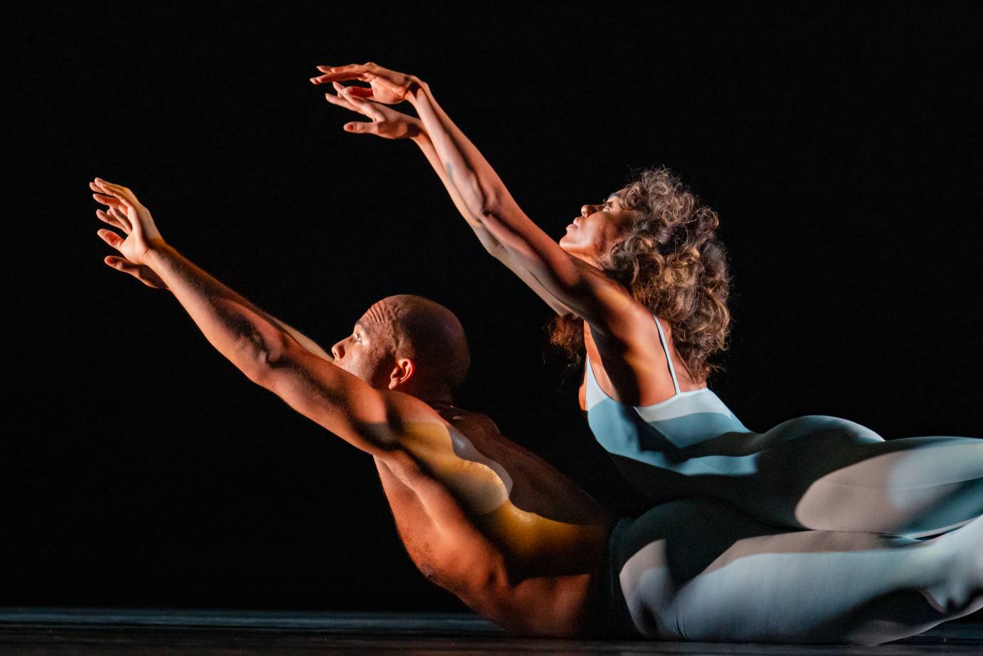 A male dancer lies on the floor, a woman dancer on top of him. Wearing variations on blue costumes (he in tights, she in a spaghetti strap leotard and tights), the dancers lift there chests and arms on the diagonal. 