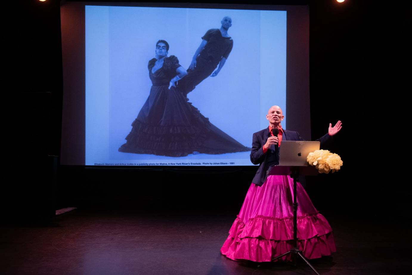 Bald headed man wearing a long, magenta flounced skirt,, magenta button down shirt and black blazer holding a microphone with a projected image of a woman in a trailing skirt with a standing man projecting from her hip
