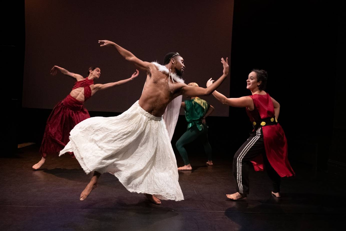 Dark-skinned man bare-chested wearing a long white skirt and tiara partners with a white women in black sweatpants striped with white and red tunic. Her dark hair is piled on her head and she is smiling. In the background a white man in a criss-crossed top and long red dress dances with a mustachioed man in blonde wig, green top and pants.
