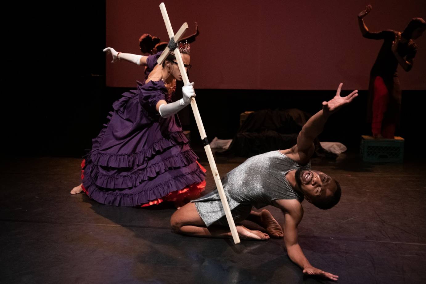 A man clothed in a long purple dress and long white gloves holds a 5' white cross as weapon against a man on his knees who kneels in front of him. The man on the ground is bent backward reaching one arm beseechingly. 