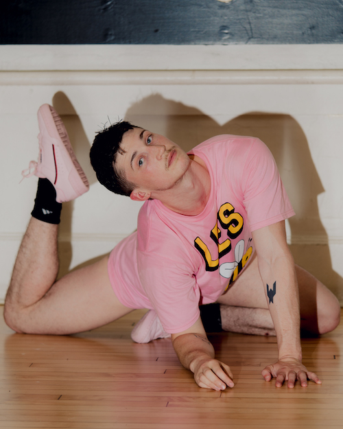 Ashley R.T. Yergens wear pink sneakers and t-shirt. He stares toward the camera while sitting on the floor. Both of his legs are bent. His right foot reaches toward his head.