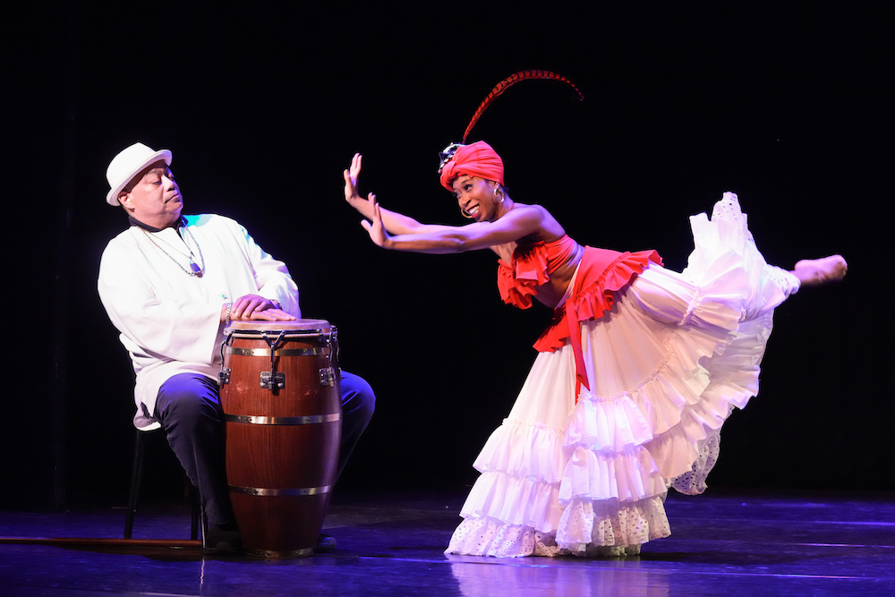 a bongo drummer rears back from Daphne Lees forward motion. His hands are on his drum ( not playing) she wearing a red turban, a red ruffled top and white ruffled skirt, Lee zestily pushes the air as if drunk, her leg extends behind her bent, sending her skirt flying.