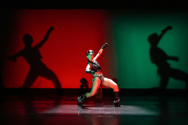 donned in motor cycle gear with studs and cap and boots Donna Clark strikes a pose looking out at the audience. She is in the center of her shadows, her silhouettes created by the lighting designer.