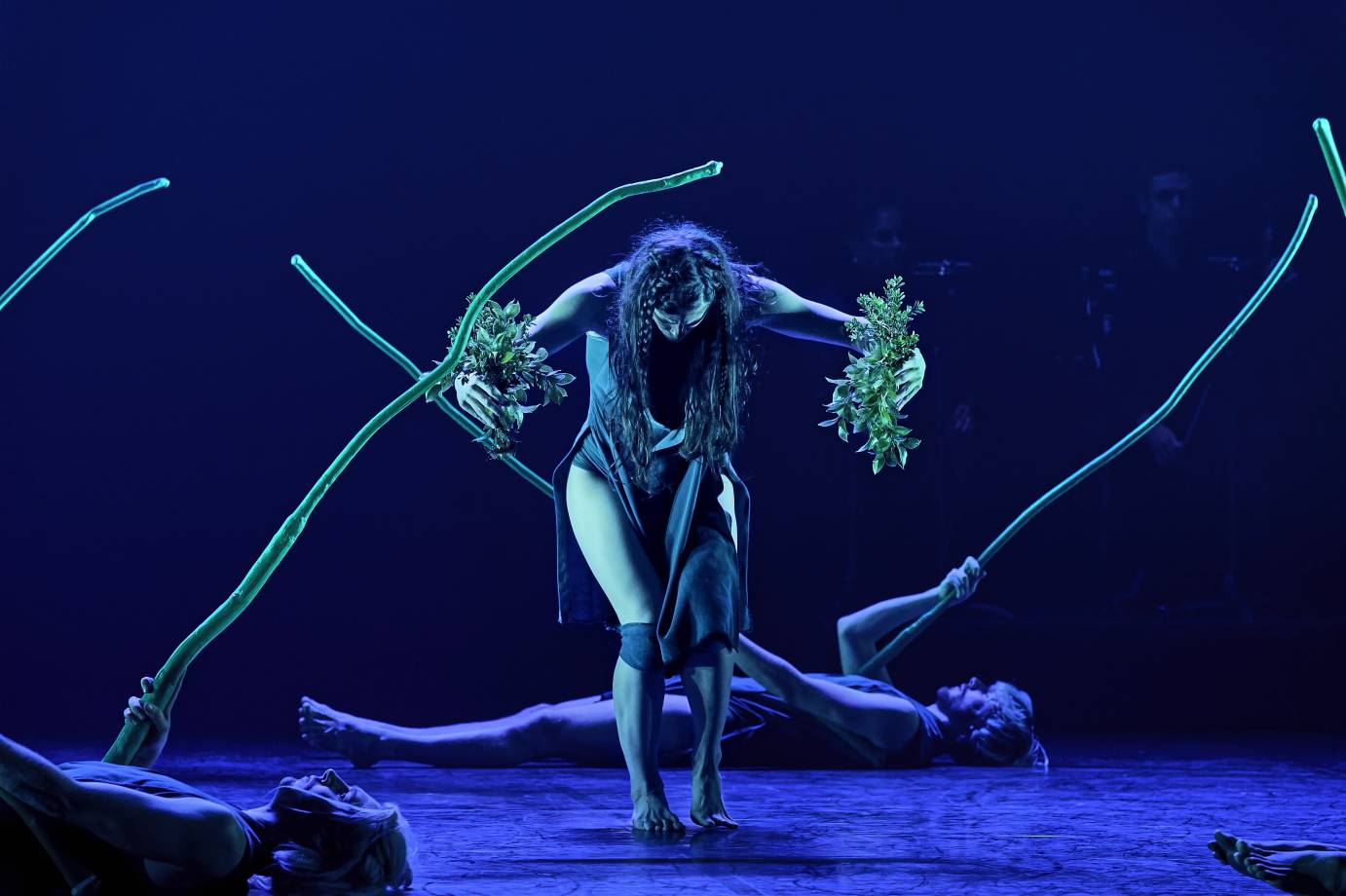 A woman with her hair in a braid holds clumps of foliage while behind her people lie on the ground with sticks 