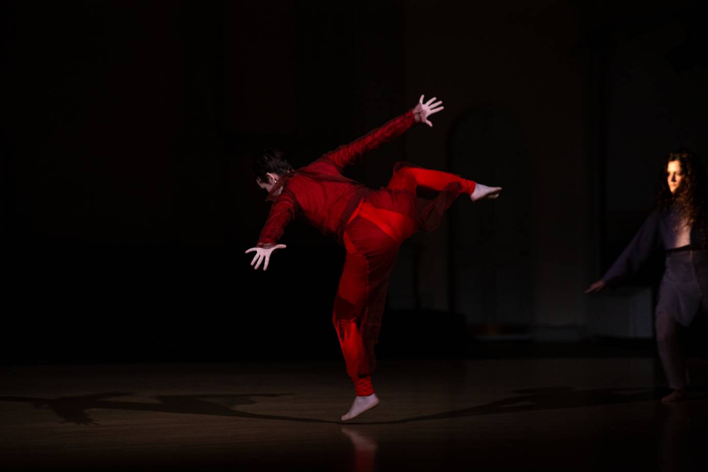 A woman in red looking to the floor in mid flight with a bent right leg, one arm reaching down and the other reaching up. Her left foot has just left the floor