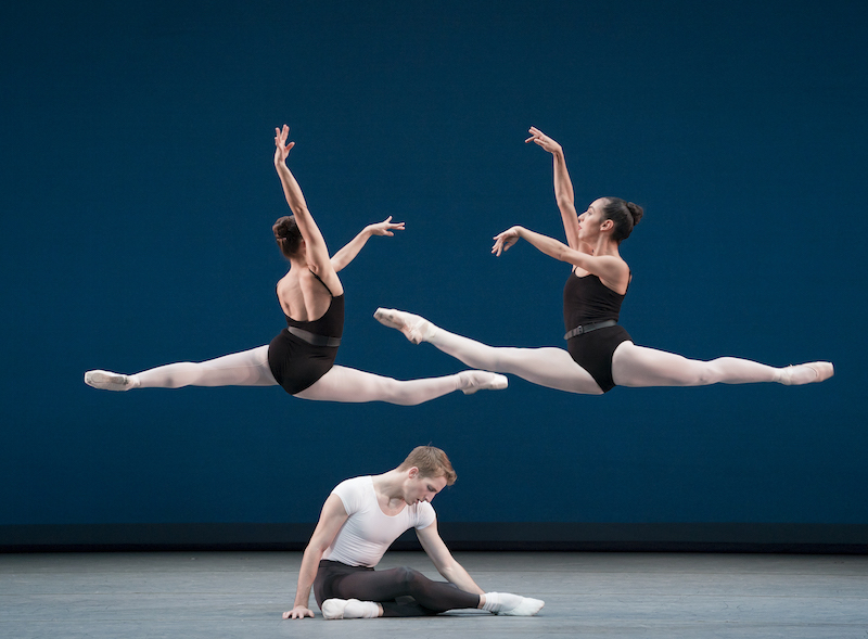 A man sits crossing his knees on the stage while two women leap above him.