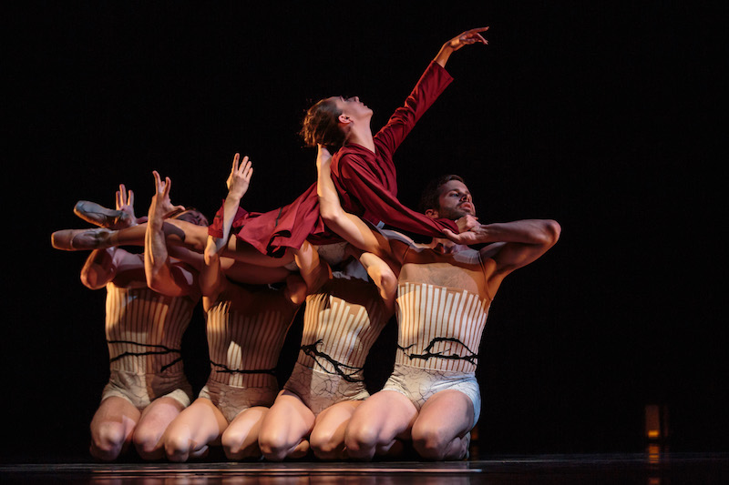 Four dancers in white unitards kneel in a line on the floor. They hold a woman in red in their arms.